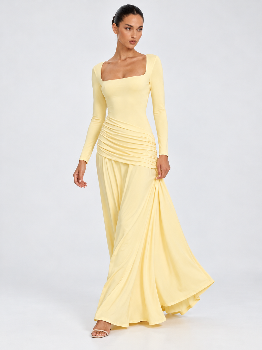 A model lifts the skirt of the Elévaison Arabella Dress in Light Yellow, showing its flowing maxi hem against a gray backdrop.