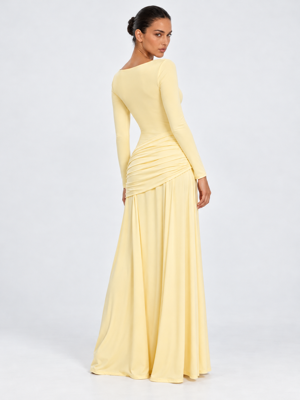 A model turns in the Elévaison Arabella Dress in Light Yellow, showing its fitted back and draped hips against a gray backdrop.