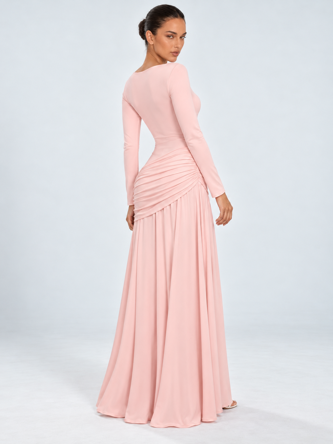 A model looks back in the Elévaison Arabella Dress in Pink, showing its fitted waist and gathered hips against a gray backdrop.