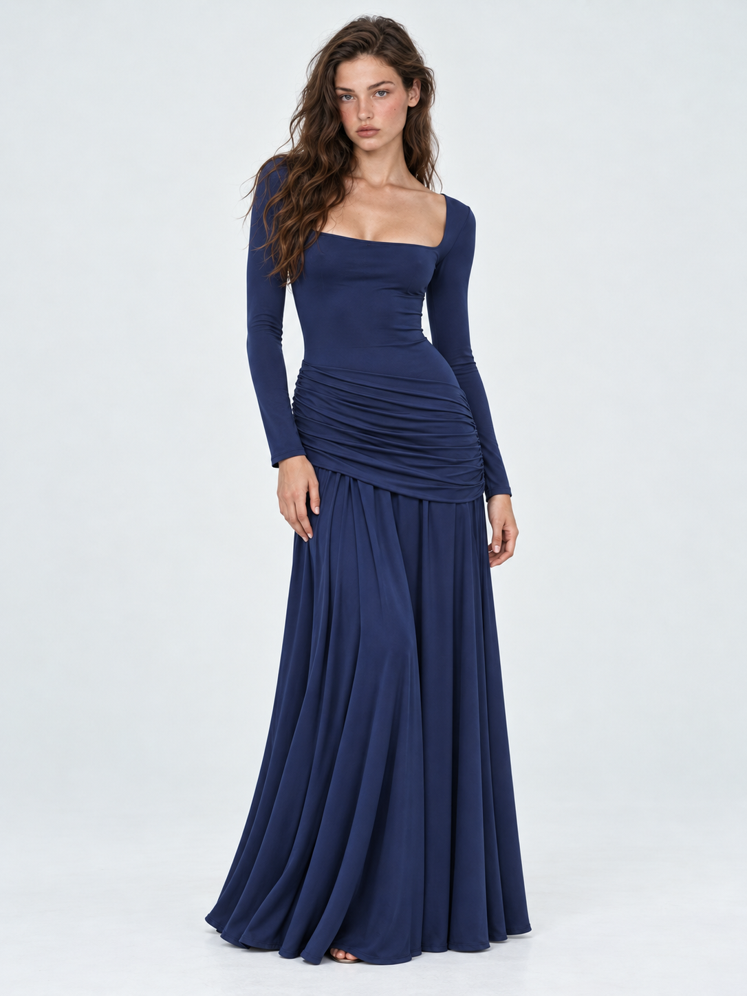 A model wears the Elévaison Arabella Dress in Dark Blue, with long sleeves and a sweeping maxi skirt against a light gray backdrop.