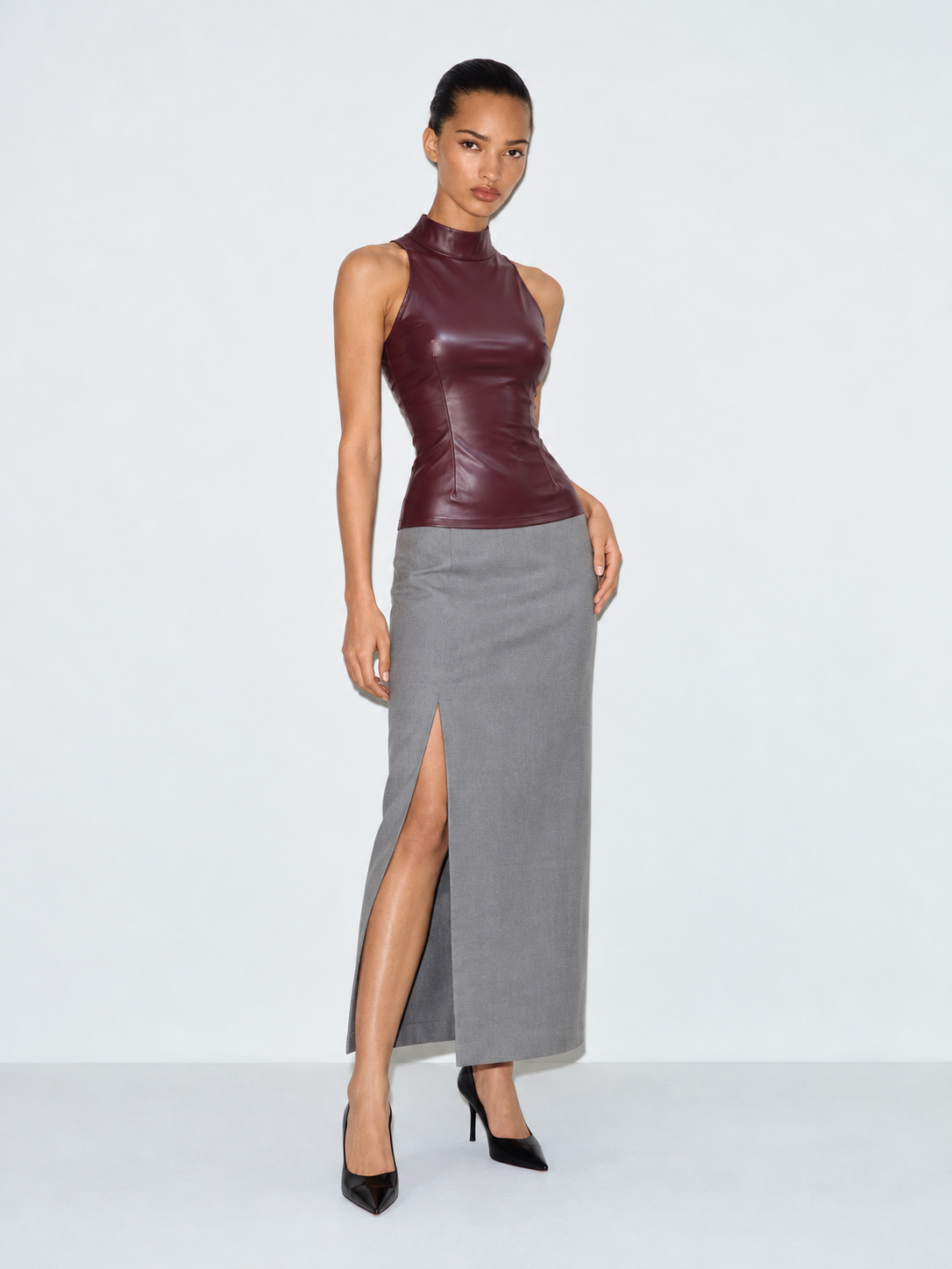 A model wears the Elévaison Carmen Top in Burgundy with a gray skirt, showing its high neck and fitted waist against a gray backdrop.