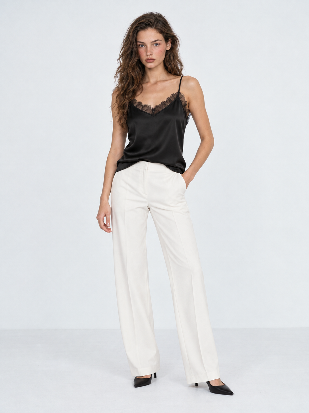 A model wears the Elévaison Anya Silk Camisole in Black with white wide-leg pants, showing its lace neckline against a gray backdrop.