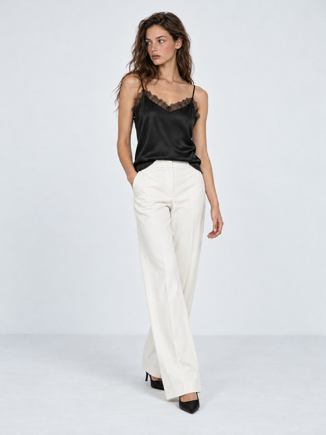 A model steps forward in the Elévaison Anya Silk Camisole in Black with white pants against a light gray studio backdrop.