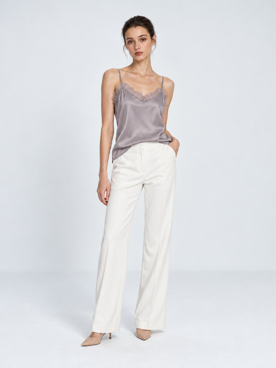 A model wears the Elévaison Anya Silk Camisole in Light Gray with white pants, showing its lace neckline against a light gray backdrop.